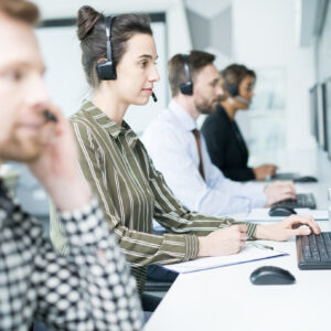 Side view portrait of several help desk operators wearing headsets sitting in row looking at computer screens and talking to clients